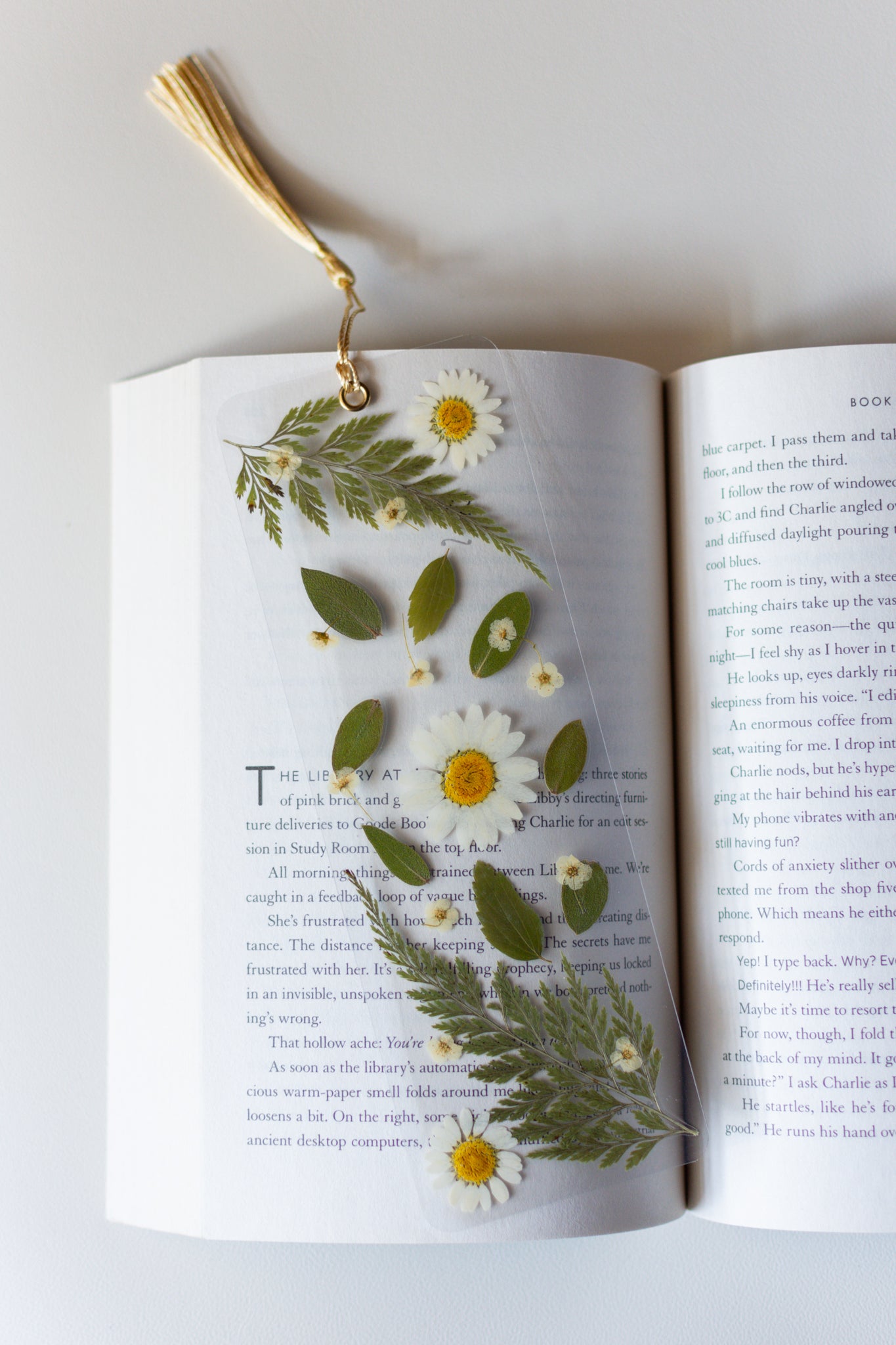 Un separador de libros transparente con flores prensadas en su interior de color blanco y hojas verdes.