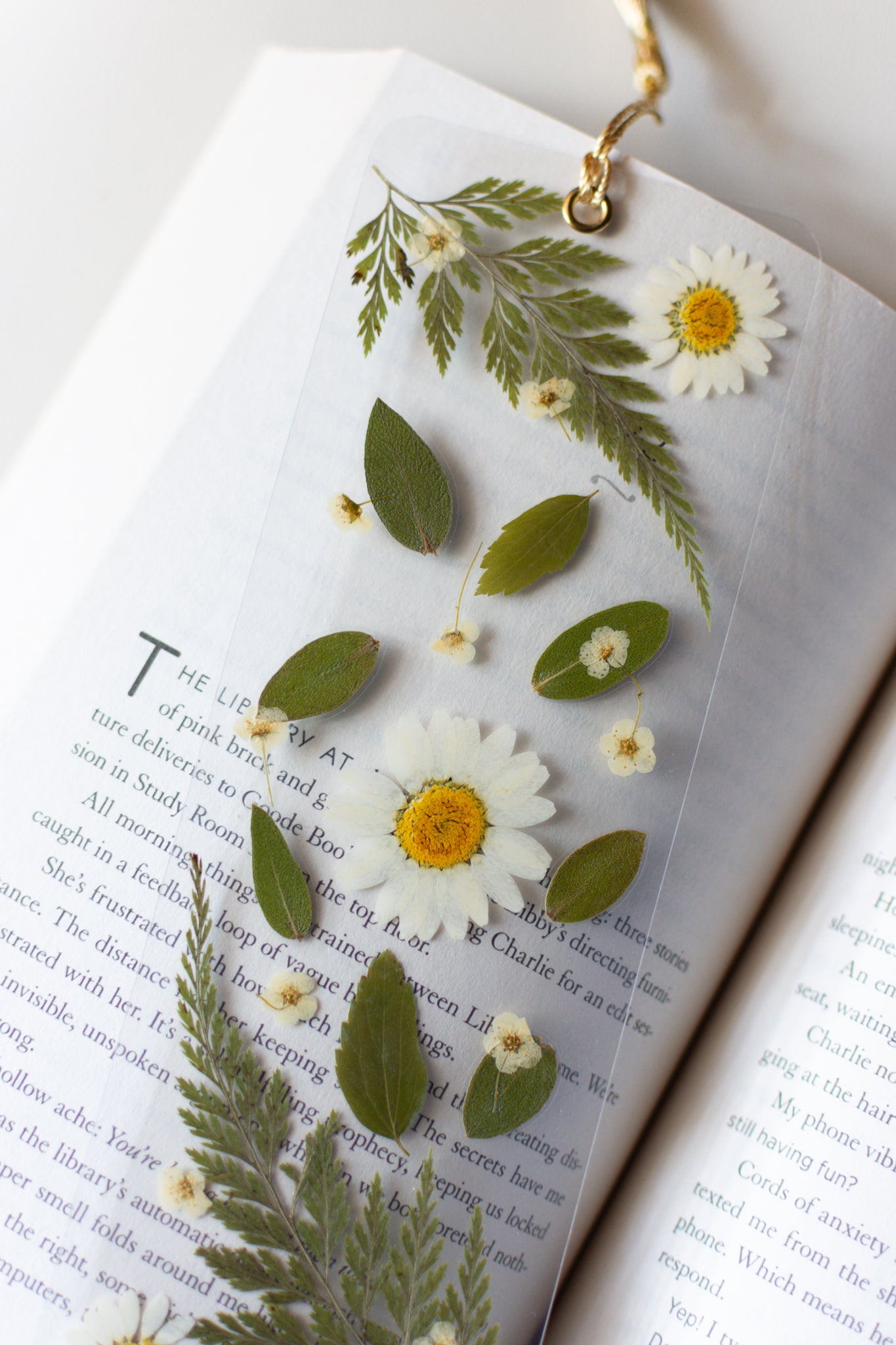 Un separador de libros transparente con flores prensadas en su interior de color blanco y hojas verdes.