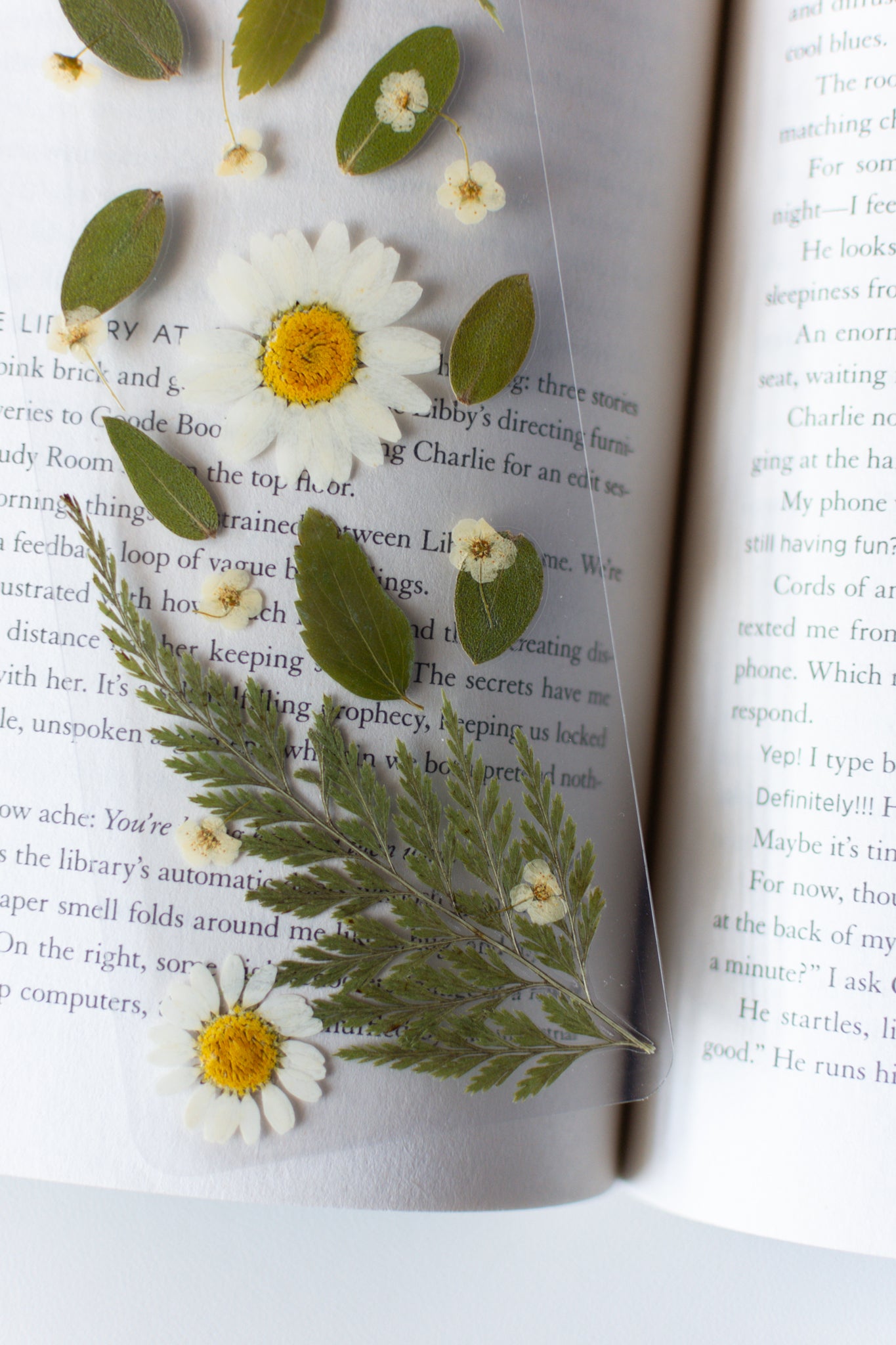 Un separador de libros transparente con flores prensadas en su interior de color blanco y hojas verdes. 