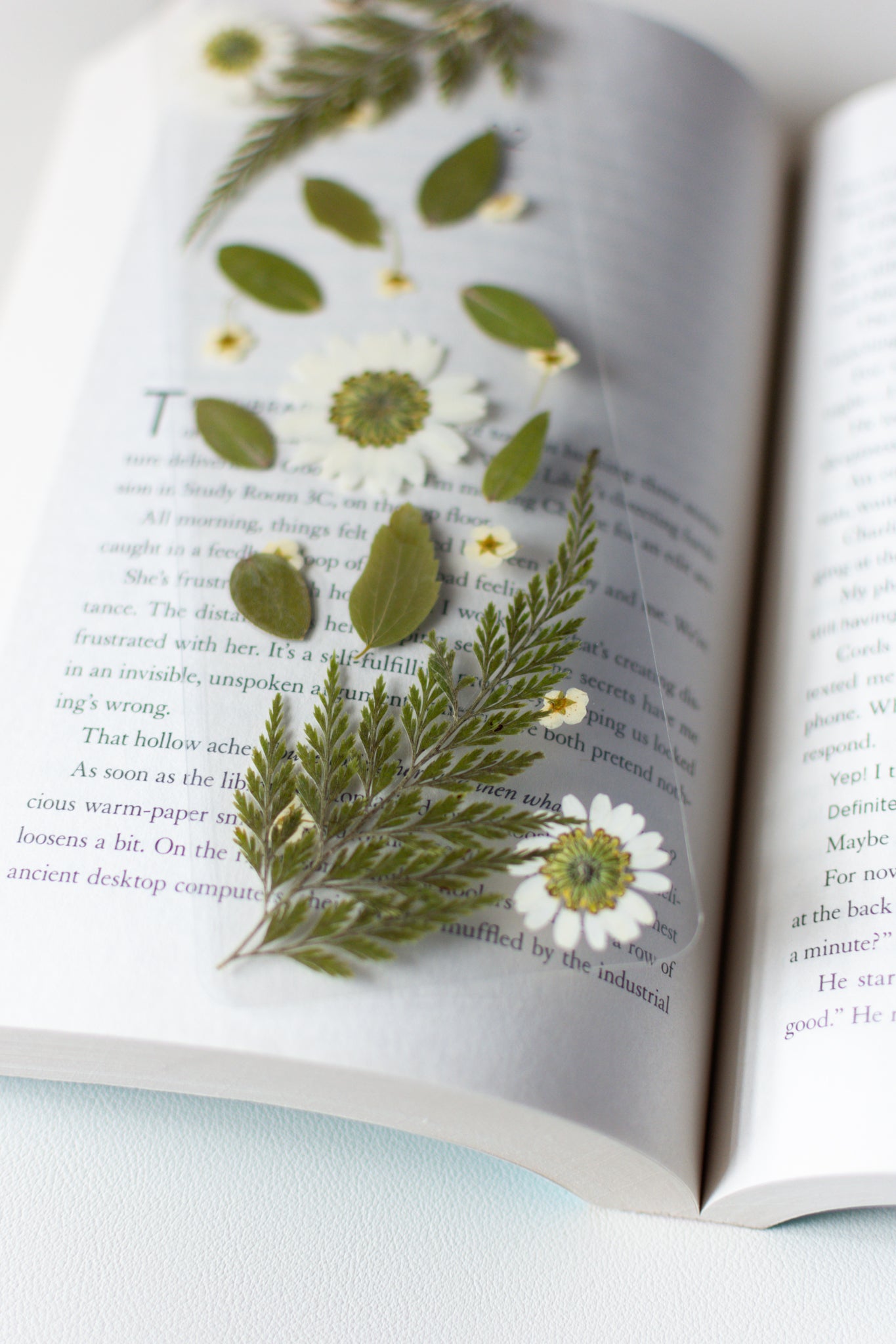 Un separador de libros transparente con flores prensadas en su interior de color blanco y hojas verdes.