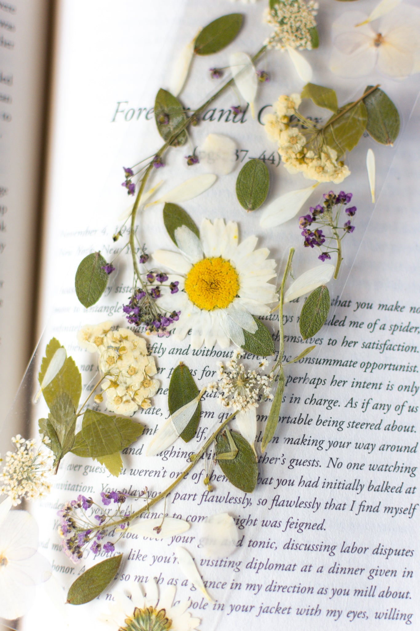 Separador de libros transparente con flores prensadas en su interior de color blanco y morado. 