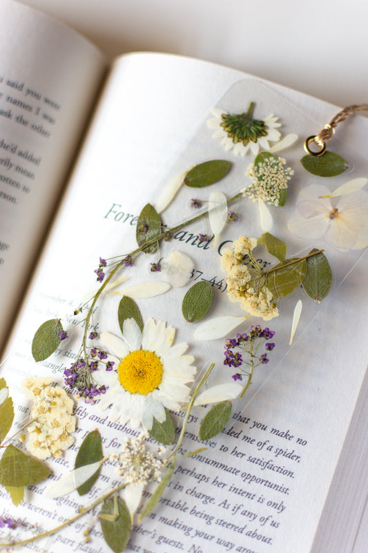 Separador de libros transparente con flores prensadas en su interior de color blanco y morado. 