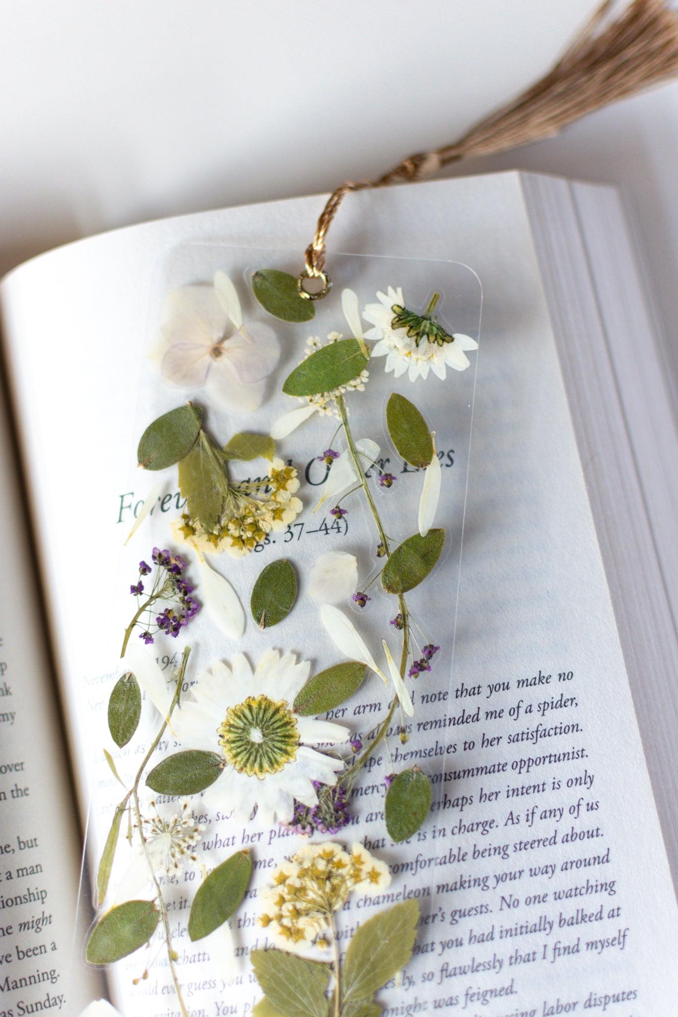 Separador de libros transparente con flores prensadas en su interior de color blanco y morado. 